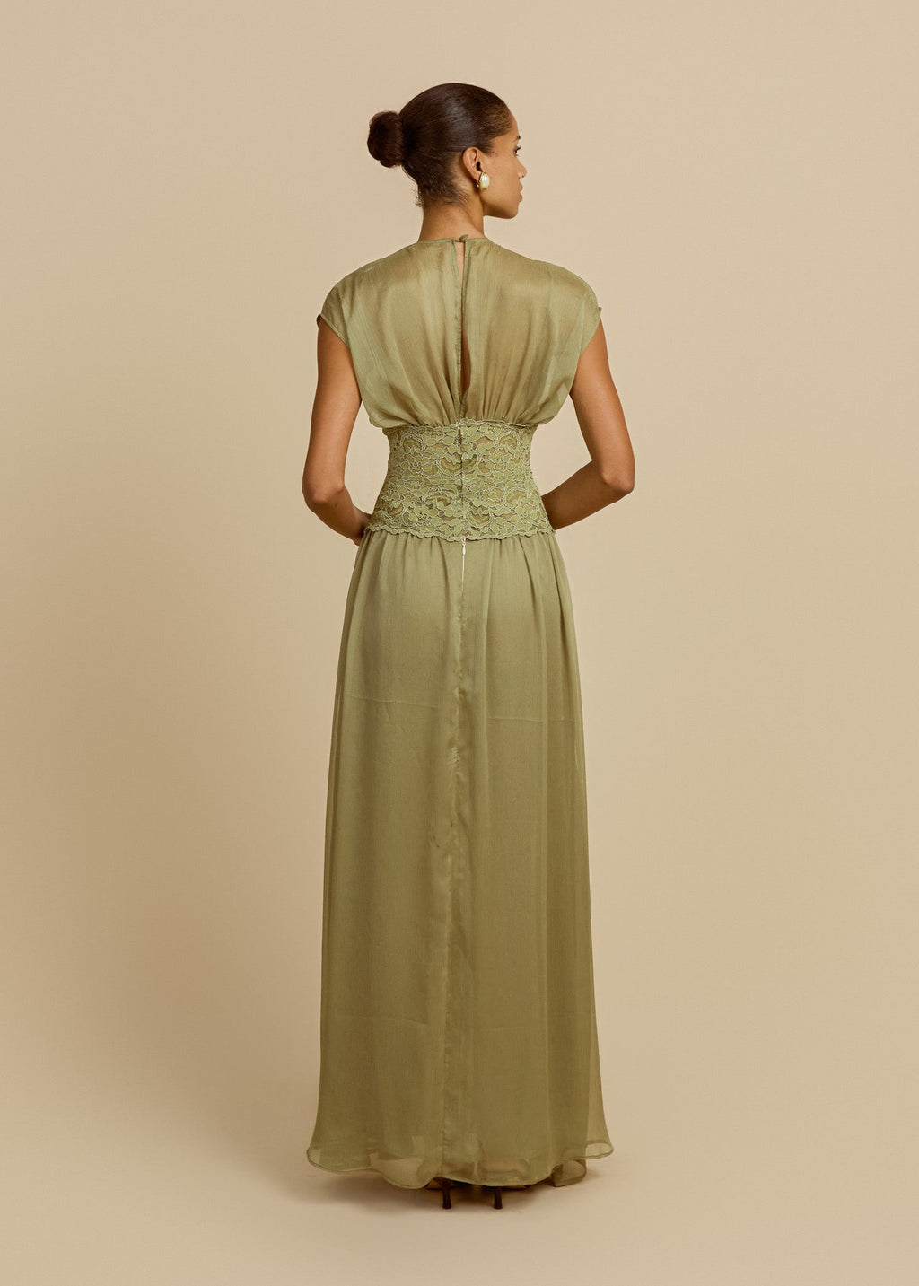 Model wearing a pistachio green floor-length dress featuring soft cap sleeves, fitted lace waist and flowing sheer chiffon skirt. Facing backwards