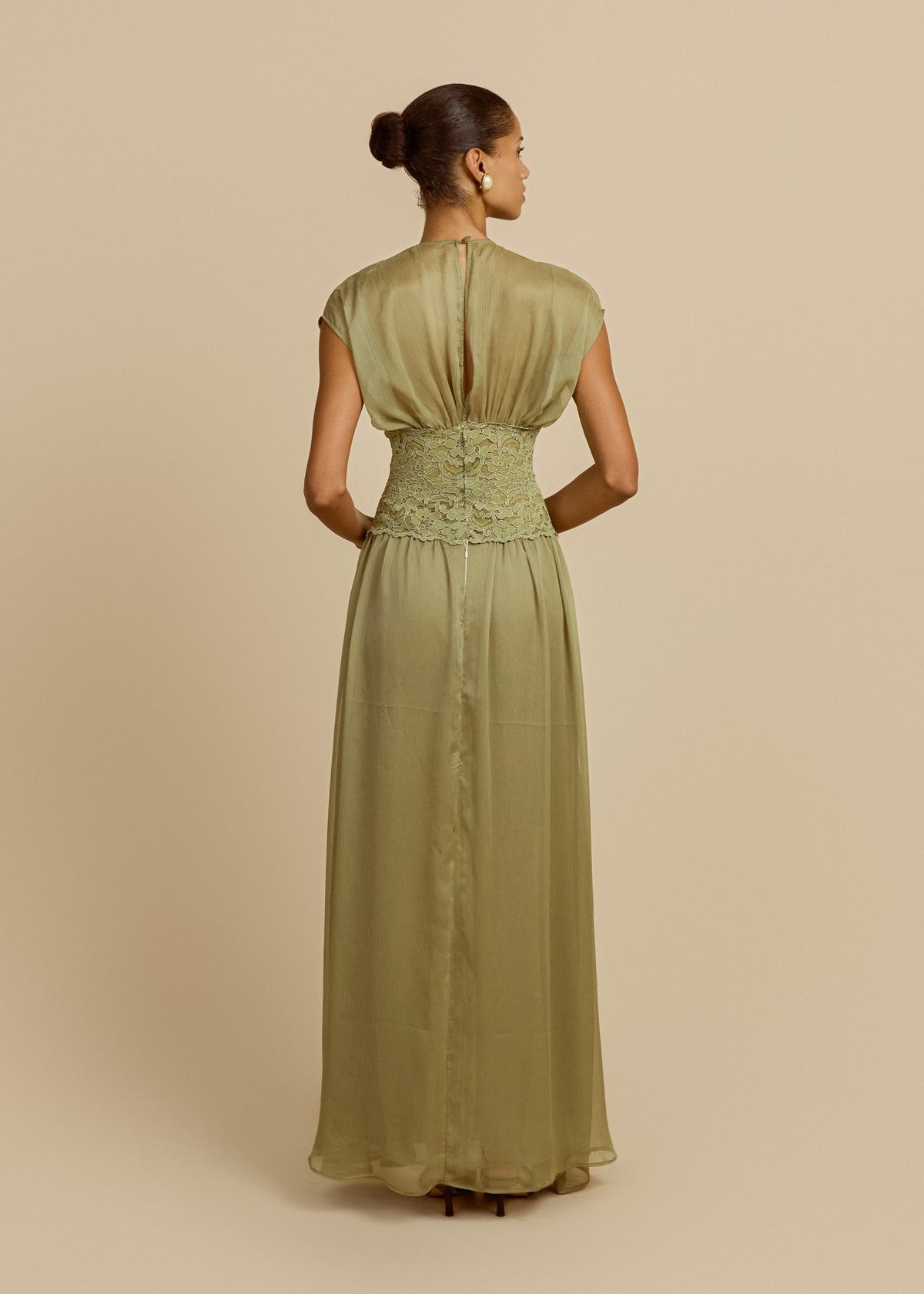 Model wearing a pistachio green floor-length dress featuring soft cap sleeves, fitted lace waist and flowing sheer chiffon skirt. Facing backwards