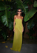 A woman wearing a strapless olive green maxi dress and black sunglasses stands in front of a backdrop of large tropical banana leaves.