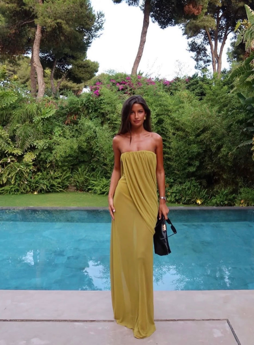 A woman stands by a swimming pool wearing a strapless olive yellow maxi dress with draped detailing. She is holding a small black handbag, with a lush green garden and trees in the background.