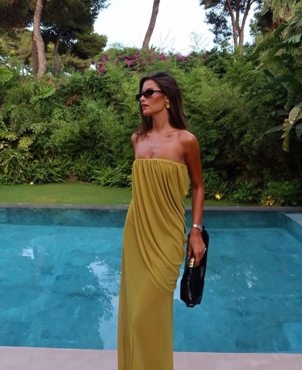 A woman stands by a swimming pool wearing a strapless olive yellow maxi dress with draped detailing. She is holding a small black handbag, with a lush green garden and trees in the background.