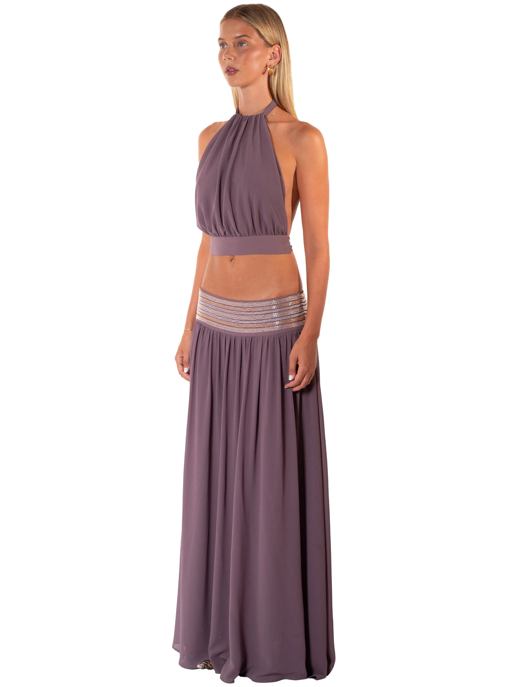 A woman modeling a two-piece dusty purple outfit consisting of a halter crop top and a matching pleated maxi skirt with a sheer mesh waistband.