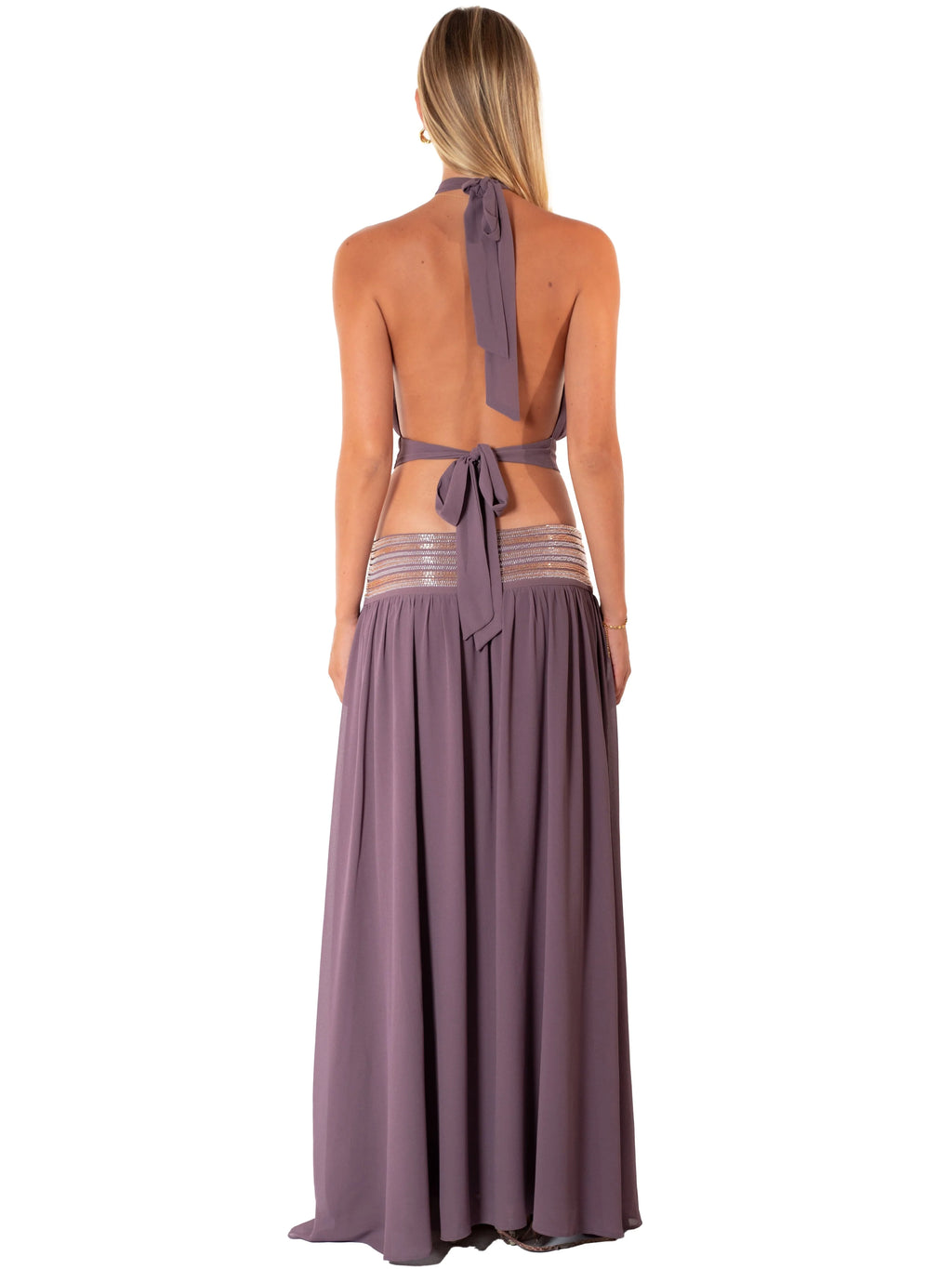 A woman shown from the back wearing a dusty purple halter maxi dress with an open back, tie detail, and sheer striped mesh panels at the waist, set against a plain white background.
