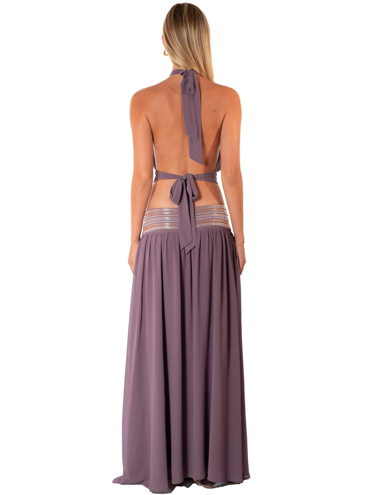 A woman shown from the back wearing a dusty purple halter maxi dress with an open back, tie detail, and sheer striped mesh panels at the waist, set against a plain white background.