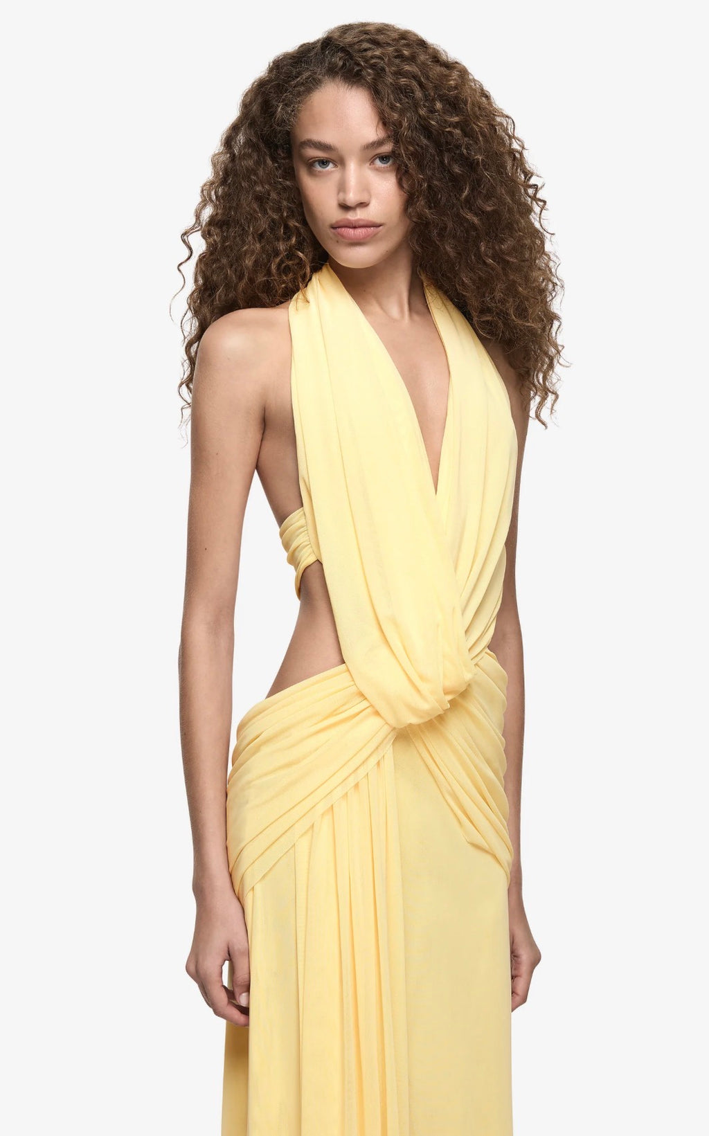A woman with long, curly brown hair wearing a pale yellow halter-neck dress with side cutouts and a draped wrap-style skirt, photographed against a plain light gray studio background.