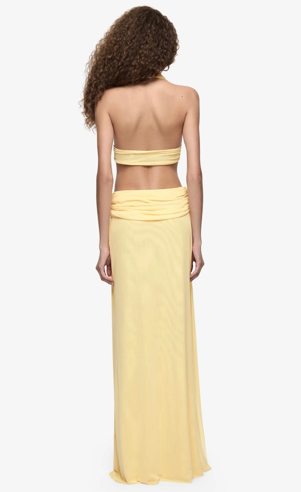 Model wearing a pale yellow halter neck maxi dress with a plunging neckline, draped waist detailing and floor-length silhouette.
