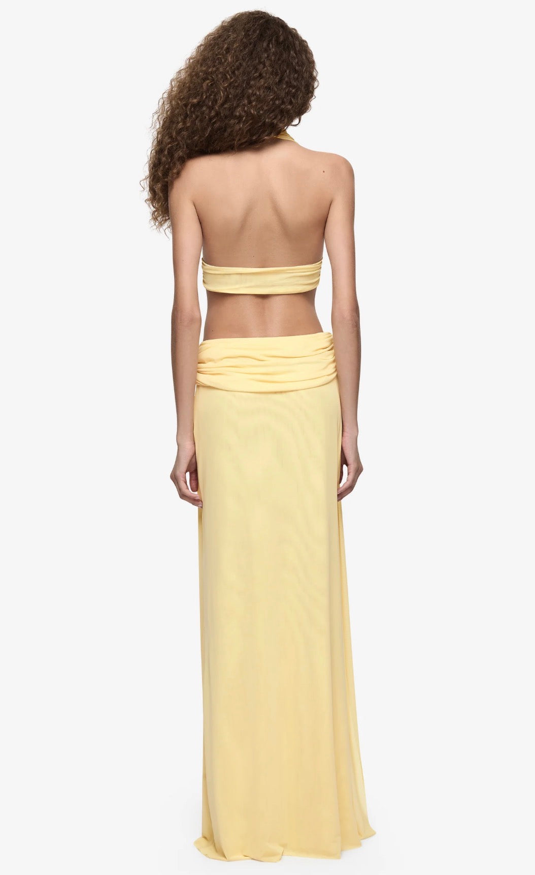  Model wearing a pale yellow halter neck maxi dress with a plunging neckline, draped waist detailing and floor-length silhouette.
