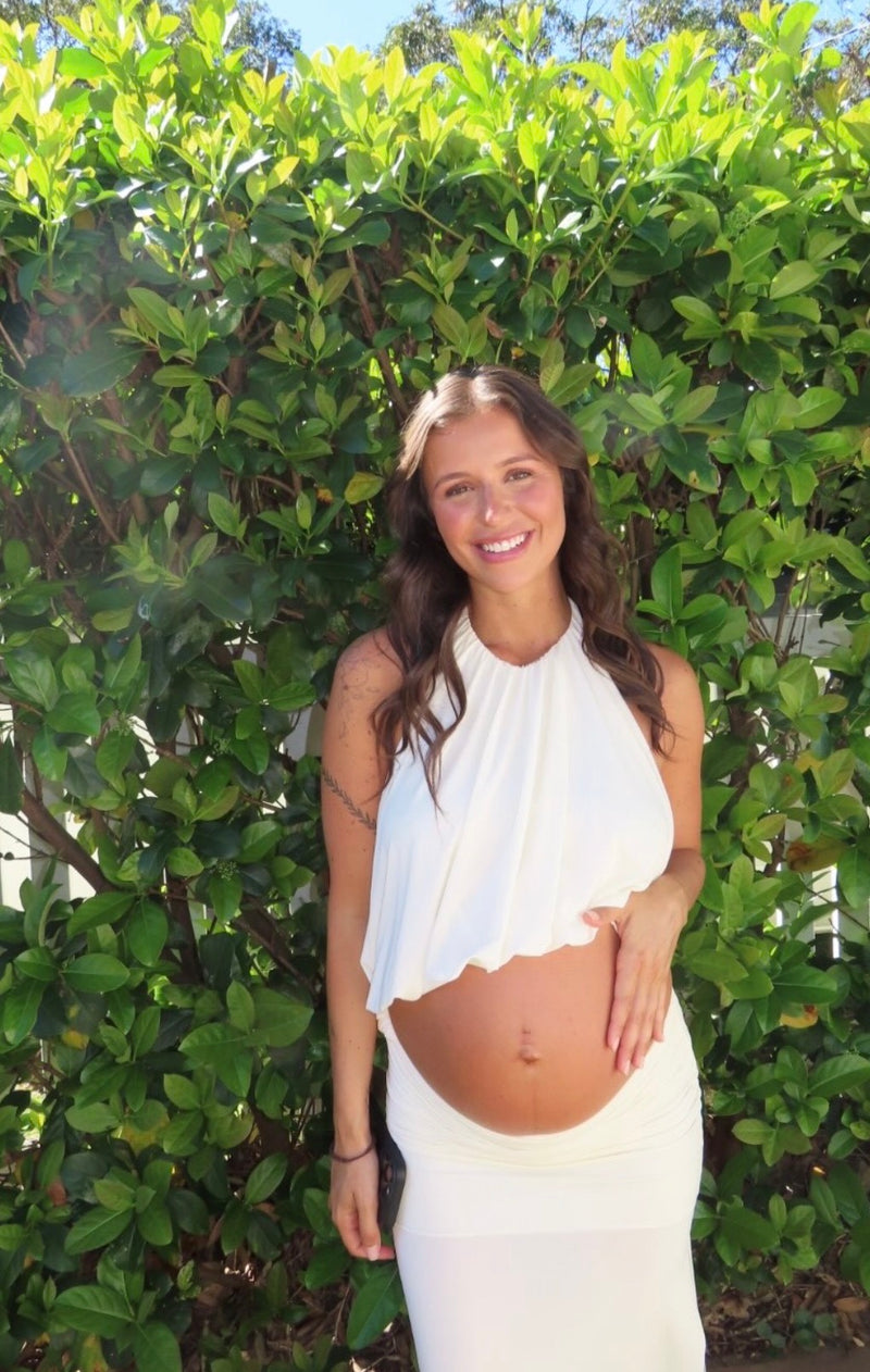 A smiling pregnant woman wearing a white sleeveless draped top and matching white skirt, standing outdoors in front of a dense green hedge. - @gracieevilla