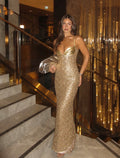 A woman wearing a sparkling gold sequined evening gown and holding a matching gold clutch, posing on a hotel lobby staircase with a decorative gold chandelier background.