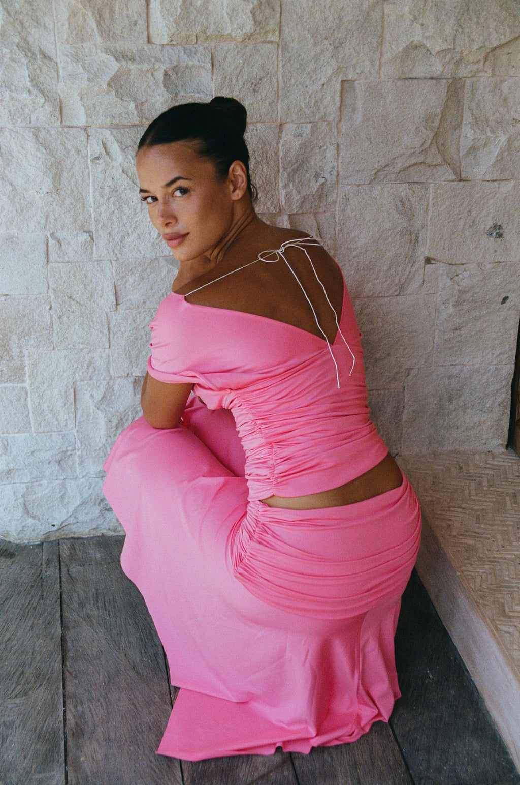 A woman wearing a bright pink off-the-shoulder maxi dress with a ruched back and tie details, posing on a stone wall and wooden floor.
