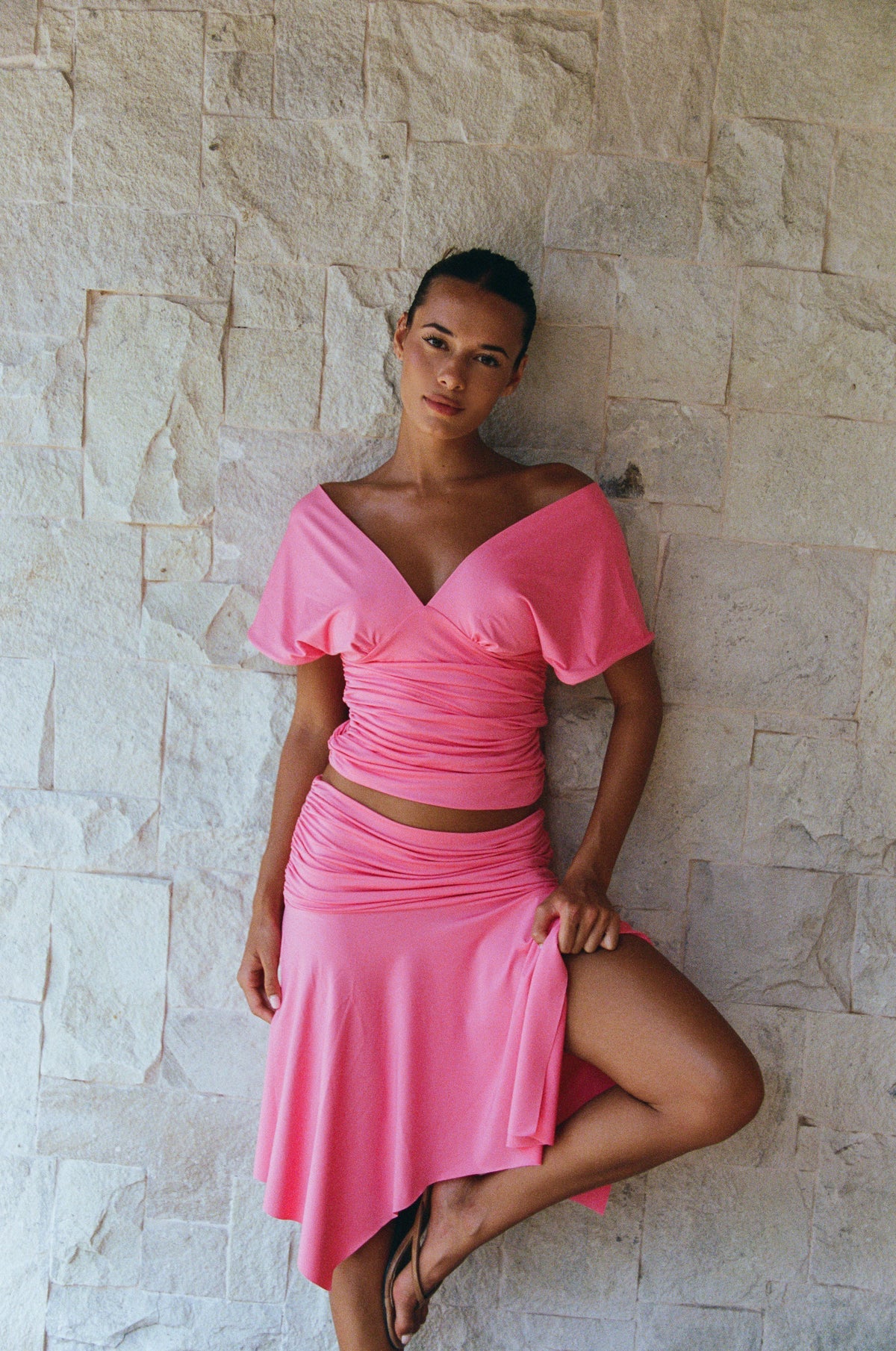 A woman posing in a bright pink two-piece set consisting of an off-the-shoulder wrap top and a matching high-waisted skirt, set against a light beige stone wall.