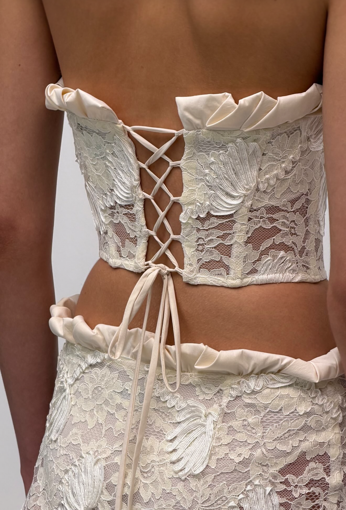 A close-up back view of a model wearing a cream lace corset-style top with ruffled trim and lace-up back detailing, shown on a phone screenshot with a 'SHOP JULLET' overlay.