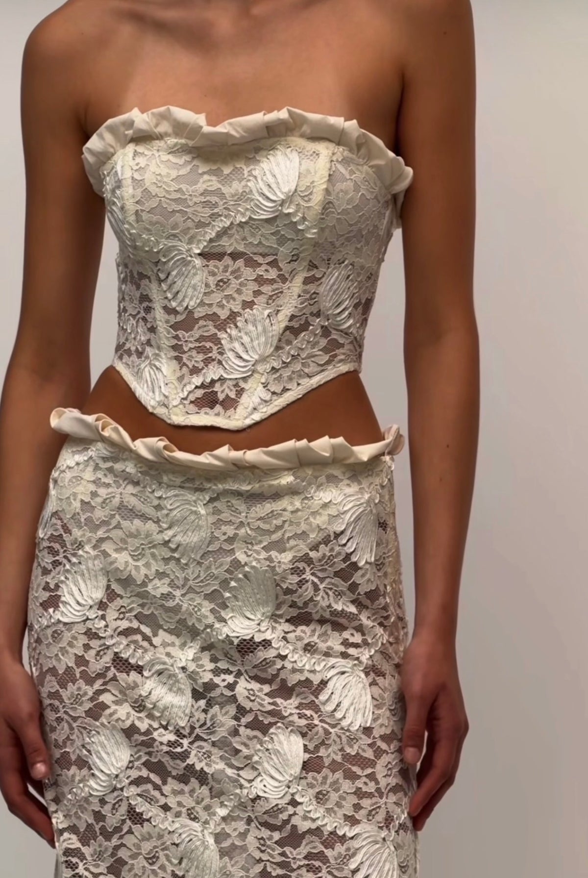 A model wearing a strapless lace two-piece outfit with a ruffled sweetheart-style top and matching ruffled high-waisted skirt, shown against a plain light background.