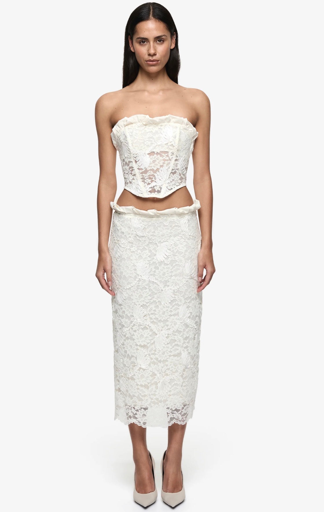 A model wearing a two-piece white lace set consisting of a strapless cropped top and a matching midi skirt, paired with beige pointed-toe heels against a plain white background.