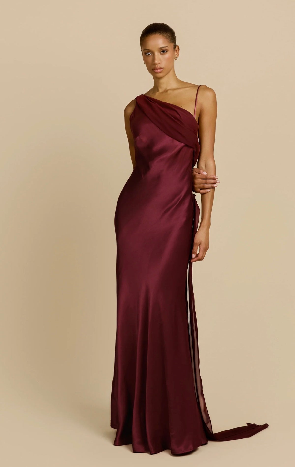 Model wearing a burgundy satin floor-length gown with an asymmetrical draped neckline, delicate strap detail and flowing silhouette against a neutral beige background.
