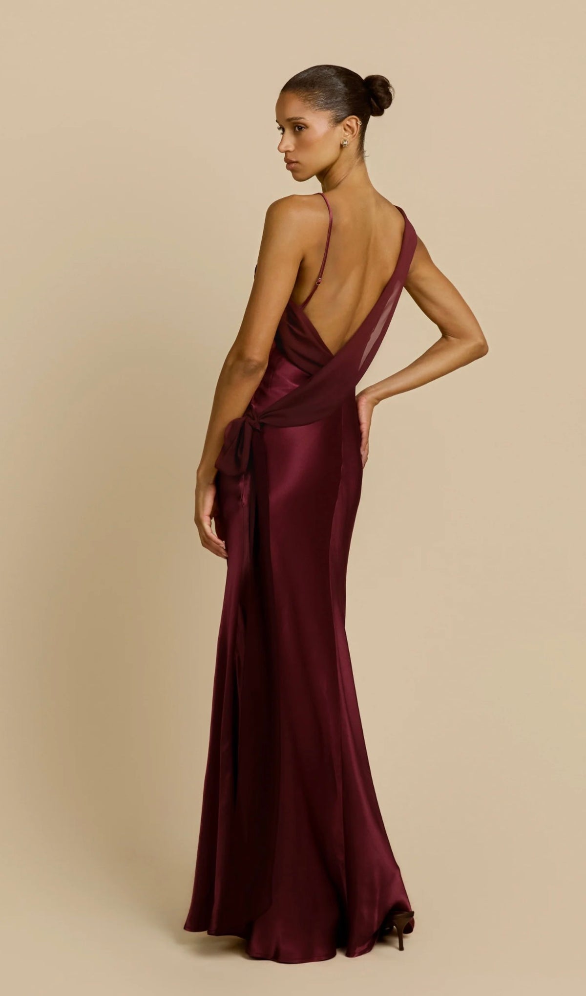 Model wearing a burgundy satin floor-length gown with an asymmetrical draped neckline, delicate strap detail and flowing silhouette against a neutral beige background.
