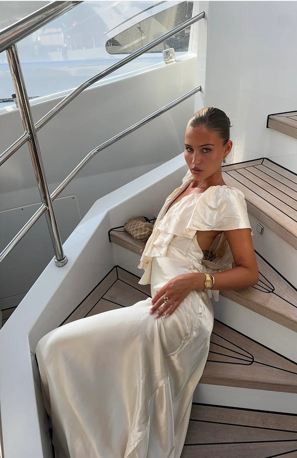 A woman wearing a cream satin-like ruffled dress sits on the wooden steps of a white yacht.