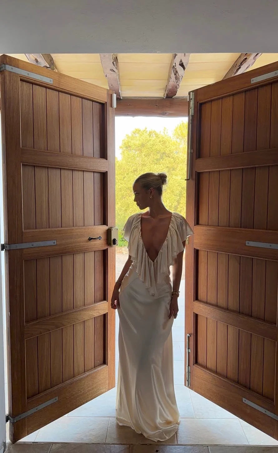 A woman wearing a sleeveless, deep V-neck, floor-length ivory dress stands in a doorway with open wooden doors, looking to the side. Bright natural light from outside illuminates the back of the dress, and green foliage is visible in the background.