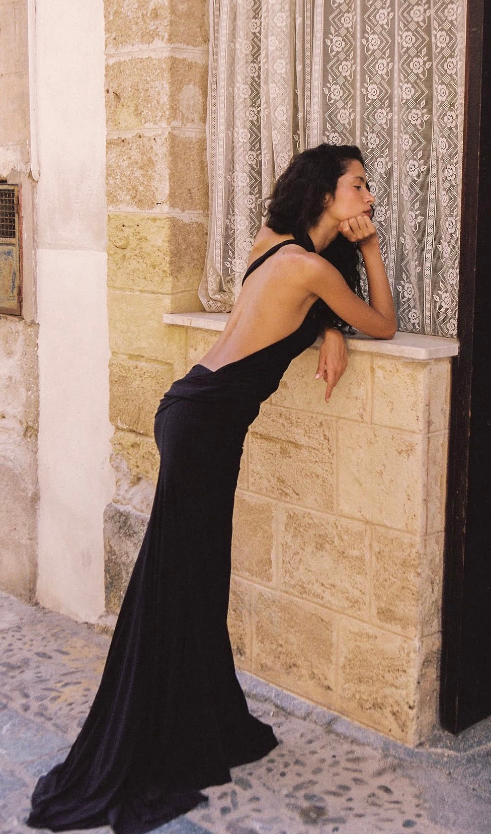  Model wearing the Natalie Rolt Odette Gown in Black, featuring an asymmetrical halter neckline, low open back, fitted silhouette and floor-length skirt with an elongated train.
