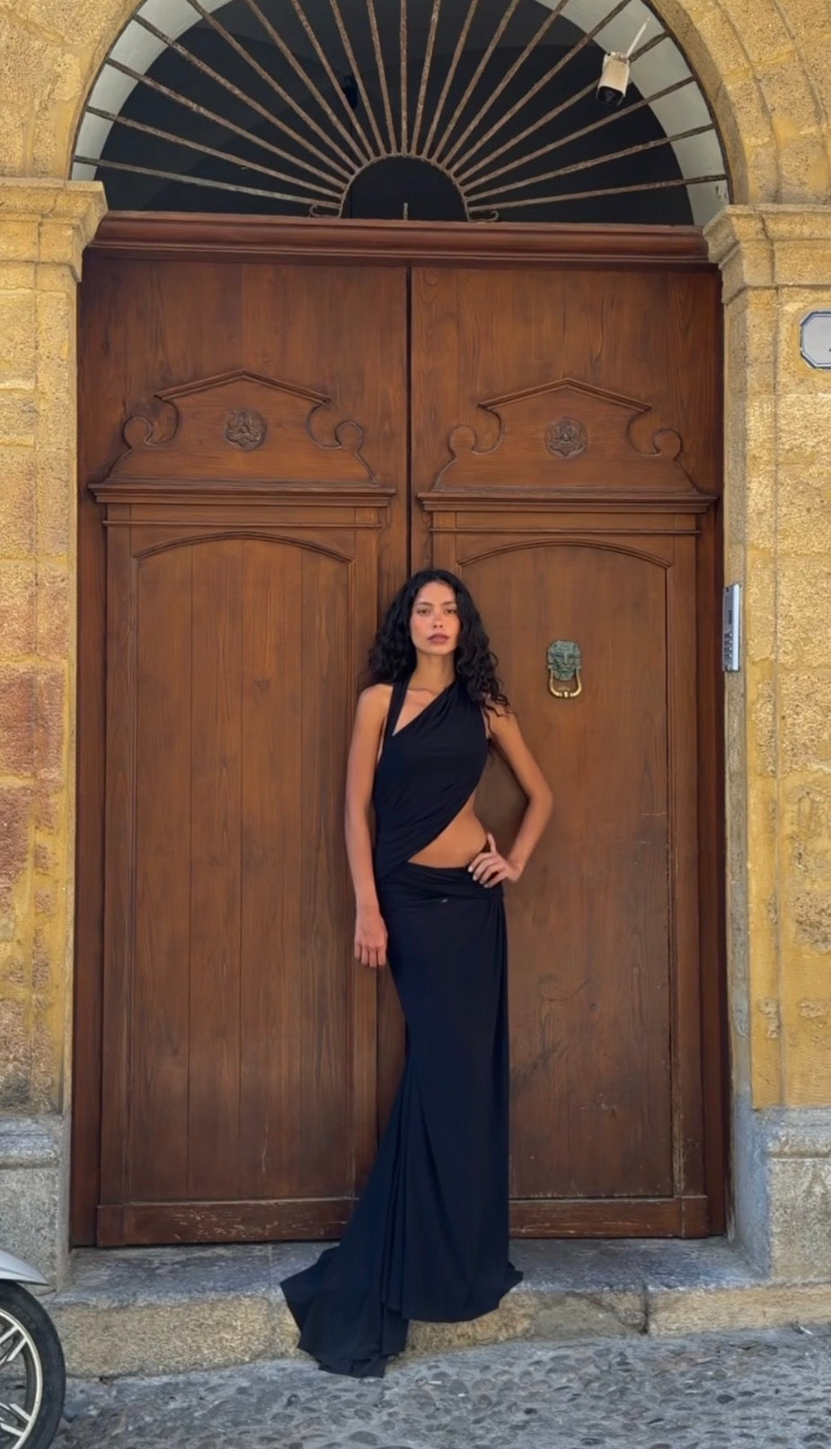 A woman wearing a black one-shoulder evening gown with a side cutout stands in front of a large, ornate wooden door set in a stone building.