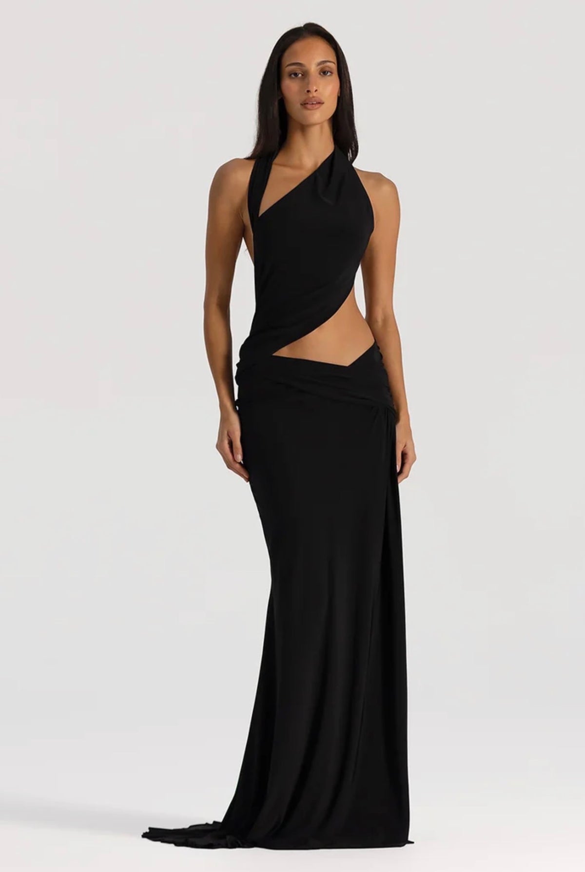 A woman modeling a floor-length black halter gown with a dramatic diagonal cutout at the waist, photographed against a plain light gray studio background.