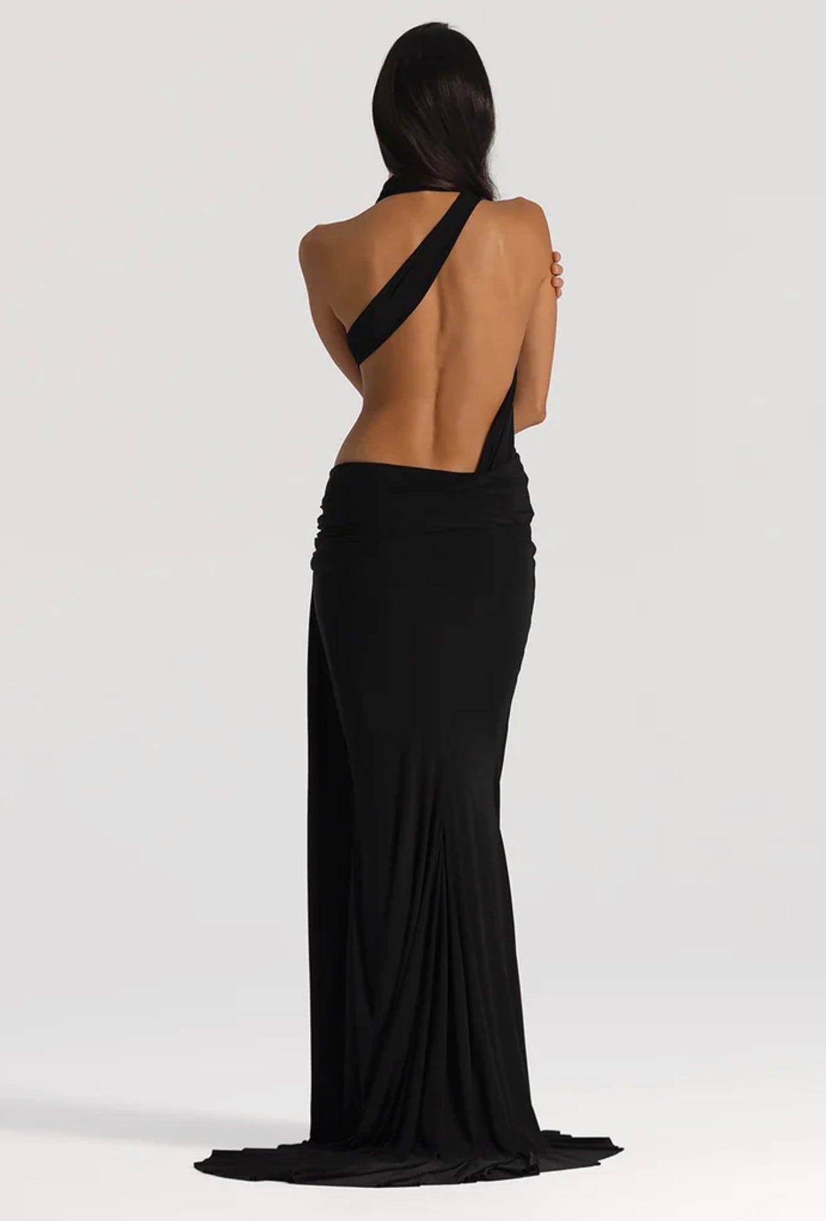 A woman shown from the back wearing a floor-length black gown with an open back and a single draped shoulder strap, photographed against a light gray studio background.