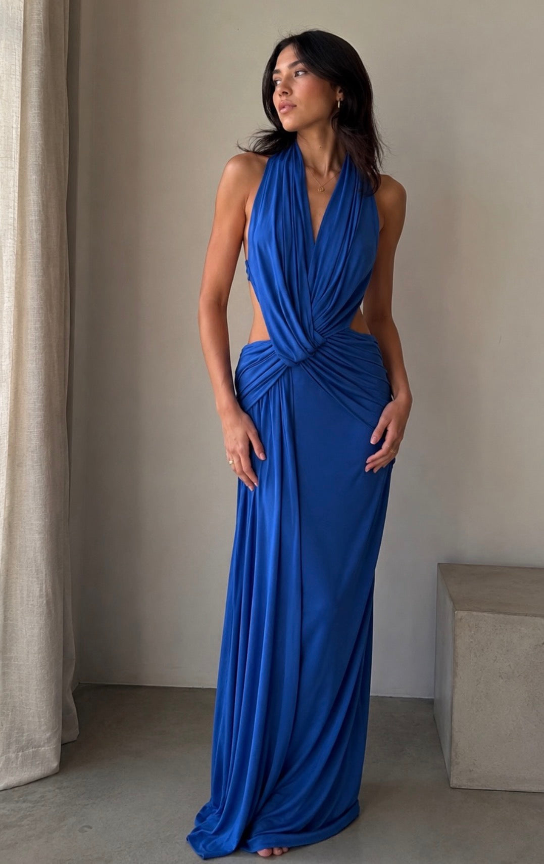 Women standing in a halter blue maxi dress infront of a beige wall.