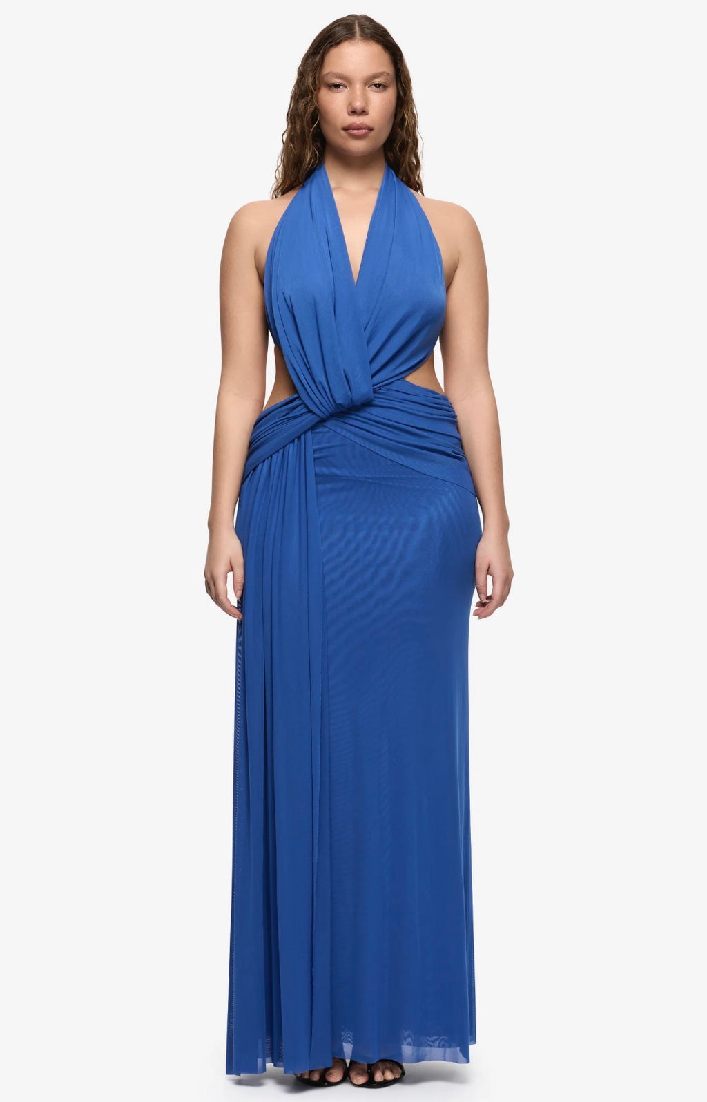 A woman modeling a royal blue halter-neck maxi dress with a twisted waist detail and side cutouts, set against a plain light gray studio background.