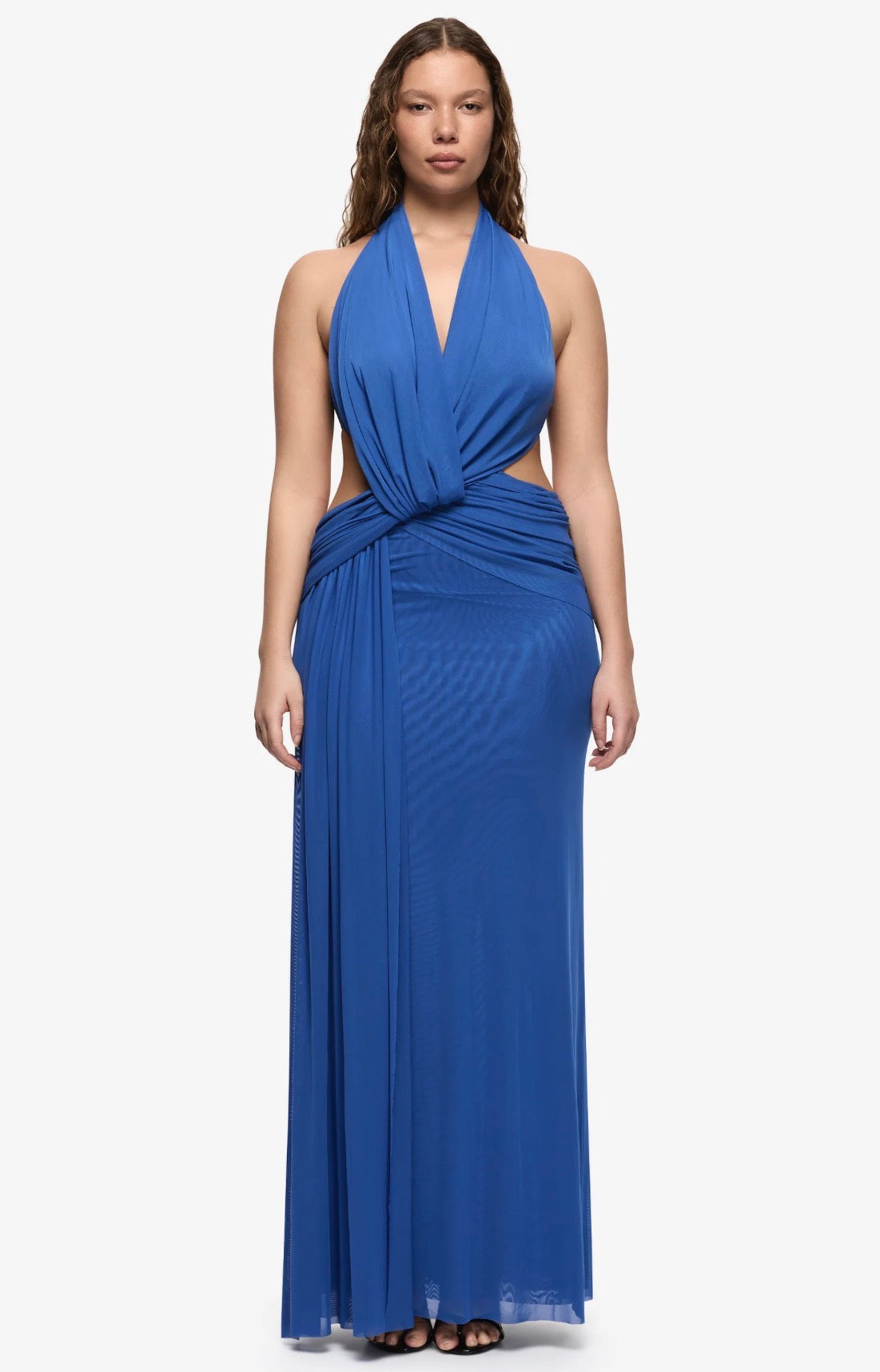 A woman modeling a royal blue halter-neck maxi dress with a twisted waist detail and side cutouts, set against a plain light gray studio background.