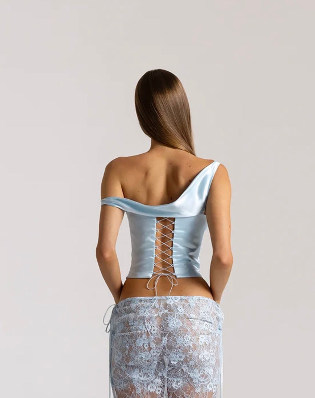 Rear view of a model wearing the Natalie Rolt Posy Top in Ice Blue, featuring an asymmetrical off-shoulder draped neckline and adjustable lace-up silk back fastening, styled with a coordinating sheer lace maxi skirt.
