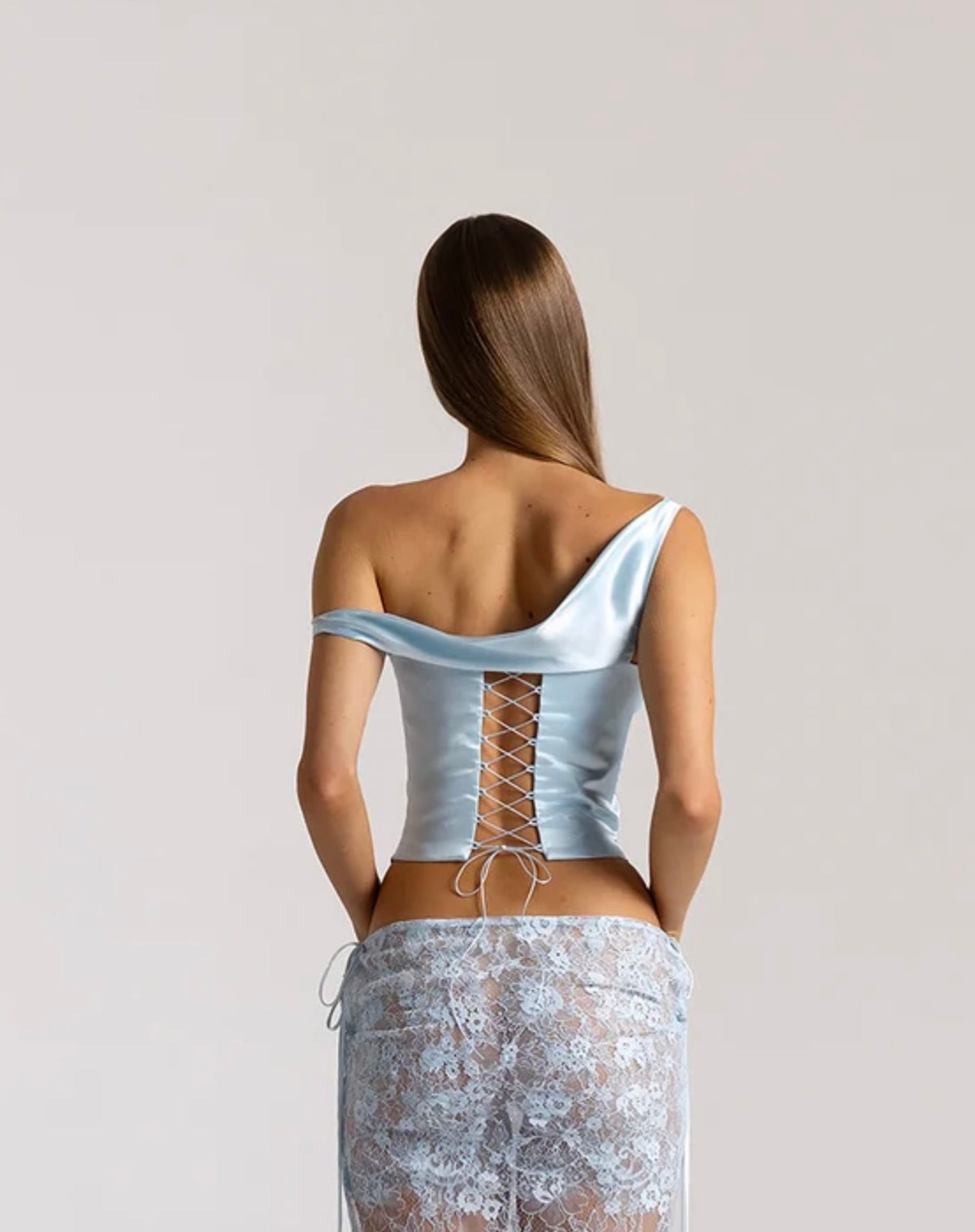 Rear view of a model wearing the Natalie Rolt Posy Top in Ice Blue, featuring an asymmetrical off-shoulder draped neckline and adjustable lace-up silk back fastening, styled with a coordinating sheer lace maxi skirt.
