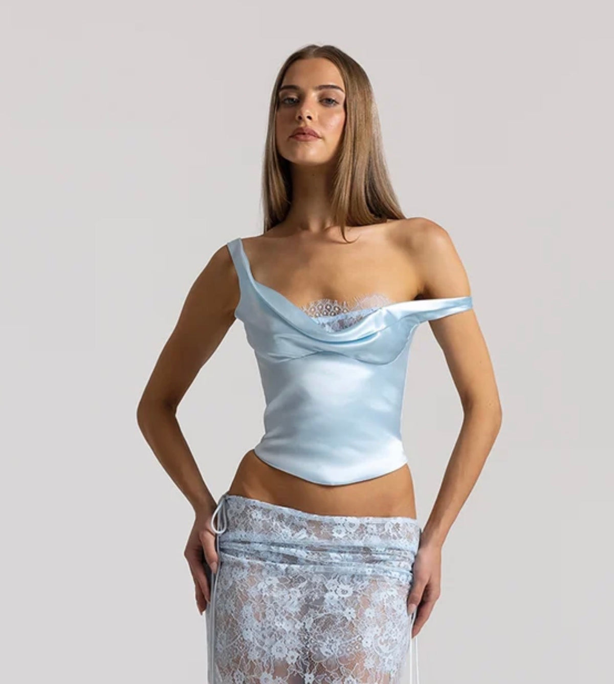Model wearing the Natalie Rolt Posy Top in Ice Blue, featuring an asymmetrical off-shoulder draped neckline with lace bust detailing and a curved hem, styled with a coordinating sheer lace maxi skirt.
