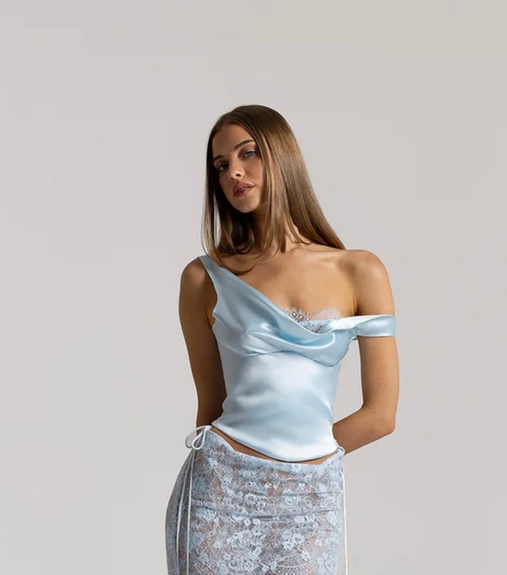 Model wearing the Natalie Rolt Posy Top in Ice Blue, featuring an asymmetrical off-shoulder draped neckline with lace bust detailing and a curved hem, styled with a coordinating sheer lace maxi skirt.
