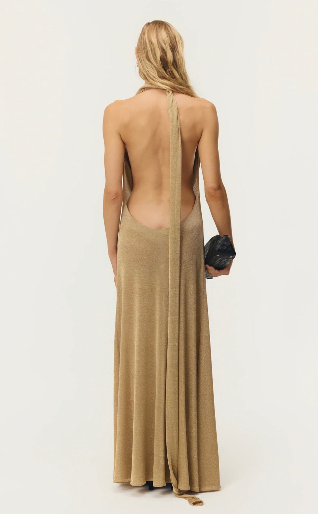 A woman shown from the back wearing a long, open-back maxi dress in a warm beige or camel shade. The dress has a single strap running down the center of the open back.
