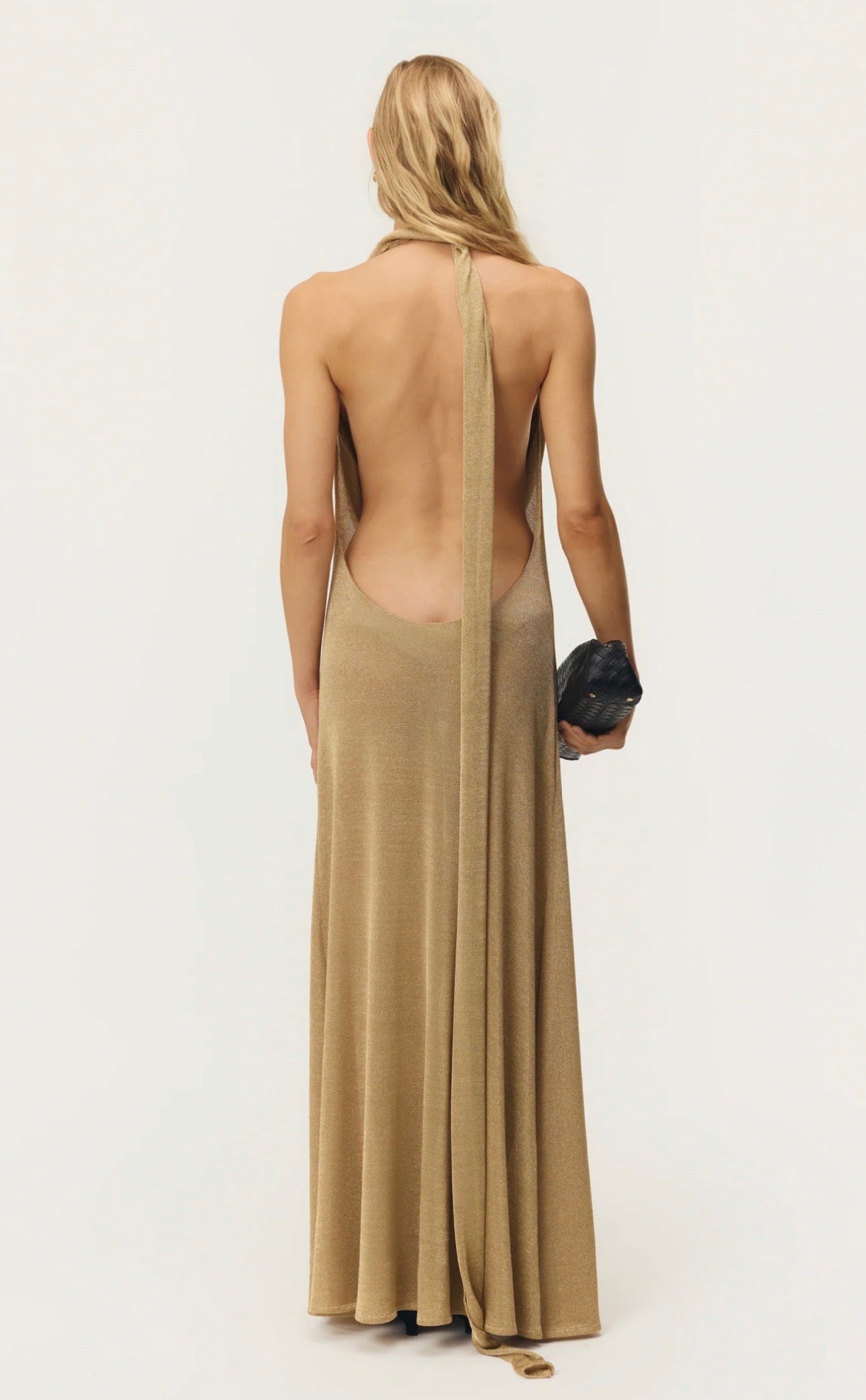 A woman shown from the back wearing a long, open-back maxi dress in a warm beige or camel shade. The dress has a single strap running down the center of the open back.