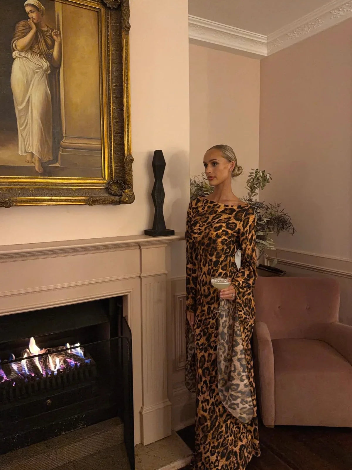  Model wearing the Helsa Rosalind leopard print maxi dress with long sleeves and draped cuff detailing, standing beside a fireplace in an elegant interior.
