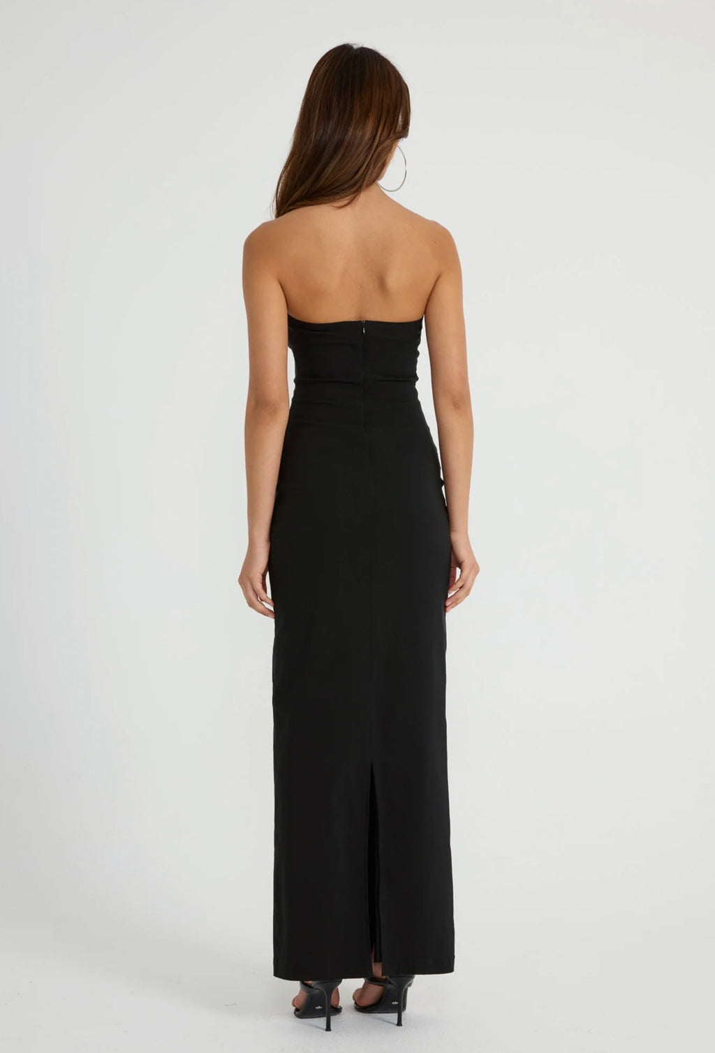 A woman viewed from the back wearing a strapless black maxi dress with a fitted bodice, side cutouts, and a front slit, standing against a plain light gray studio background.
