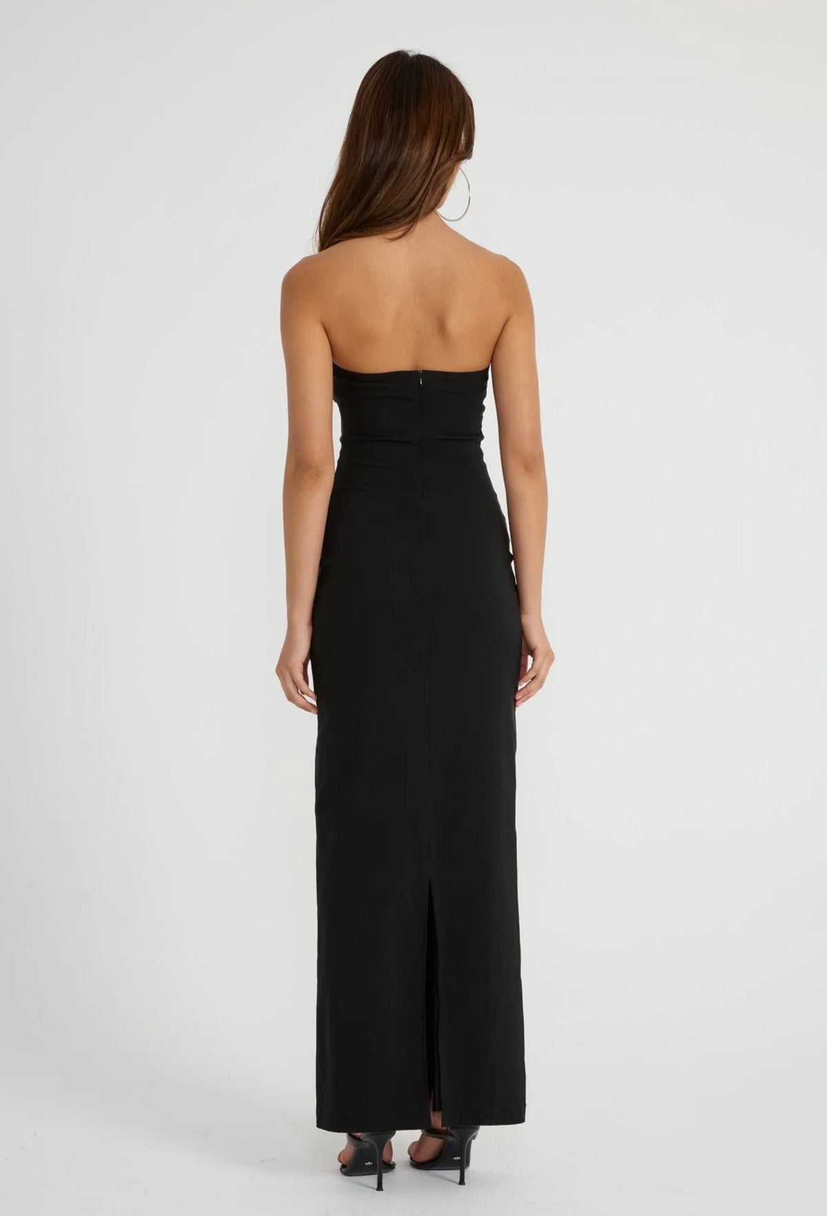 A woman viewed from the back wearing a strapless black maxi dress with a fitted bodice, side cutouts, and a front slit, standing against a plain light gray studio background.