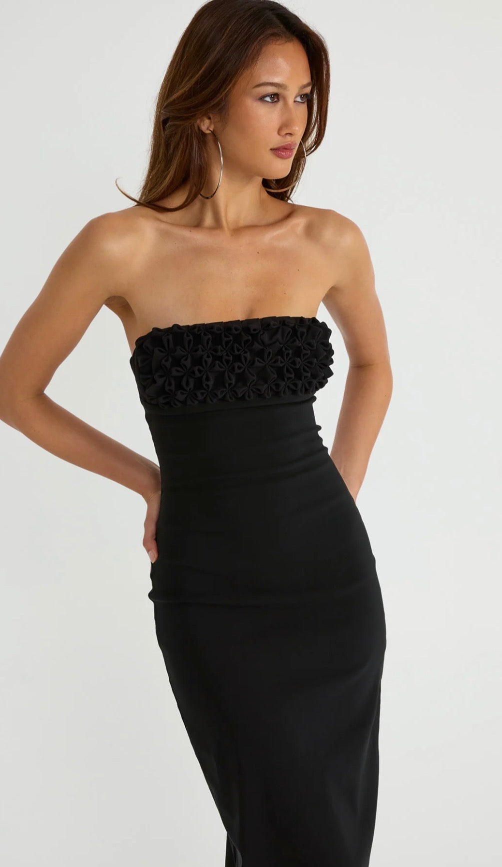A woman modeling a strapless black midi dress with a textured, ruffled strapless bodice and a fitted silhouette, set against a plain light gray studio background.