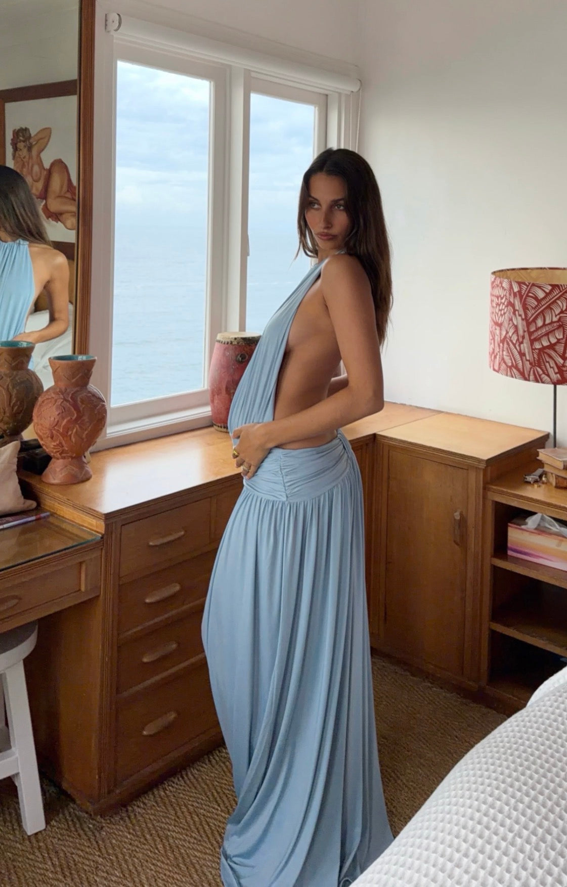 A woman wearing a light blue draped maxi dress with a low open back, standing in a bedroom with a window overlooking the sea.