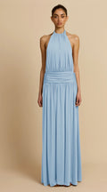 A model wearing a light blue halter-neck maxi dress with a gathered waist, standing against a solid beige studio background.