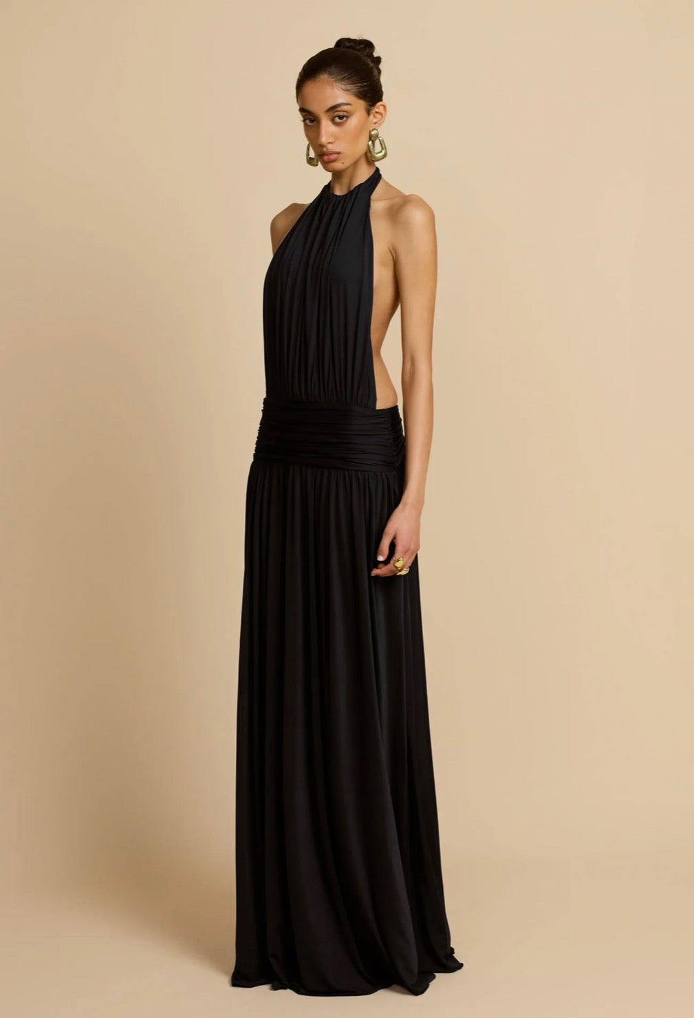 A woman modeling a floor-length black halter gown with a side cutout and gathered waist, photographed against a plain warm beige studio background.