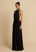 A woman modeling a floor-length black halter gown with a side cutout and gathered waist, photographed against a plain warm beige studio background.