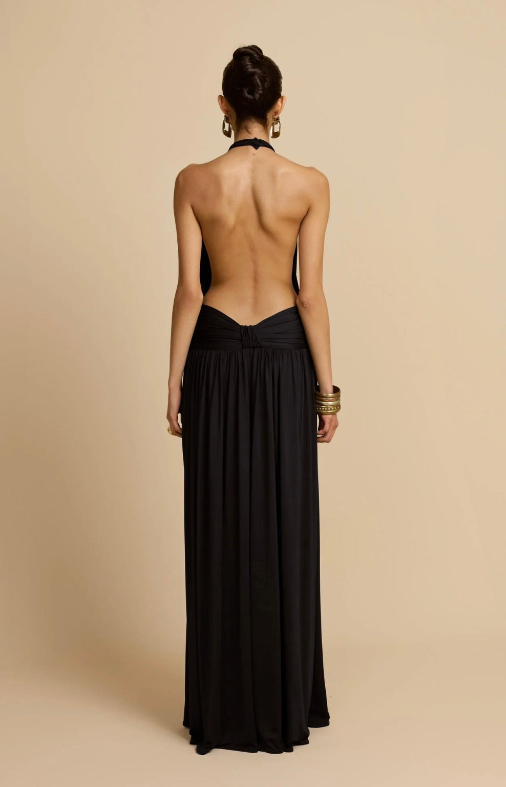 A woman shown from the back wearing a black halter-neck maxi dress with a draped open back, standing against a solid beige studio background.