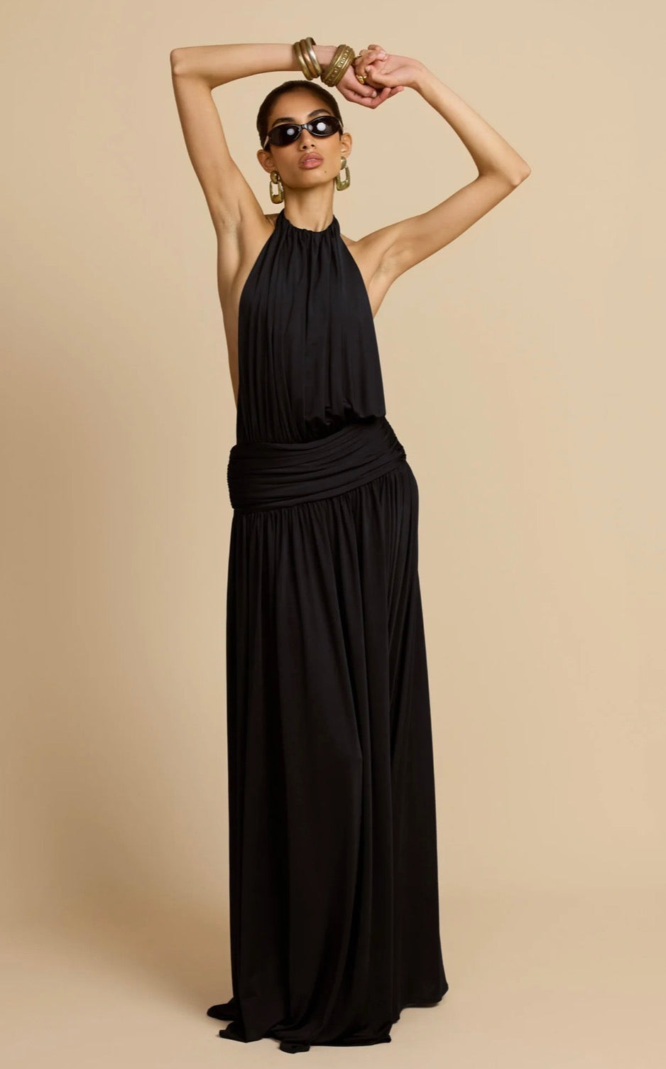 A model wearing a black halter-neck maxi dress with a draped waist, styled with gold hoop earrings and bracelets, posing against a solid beige studio background.