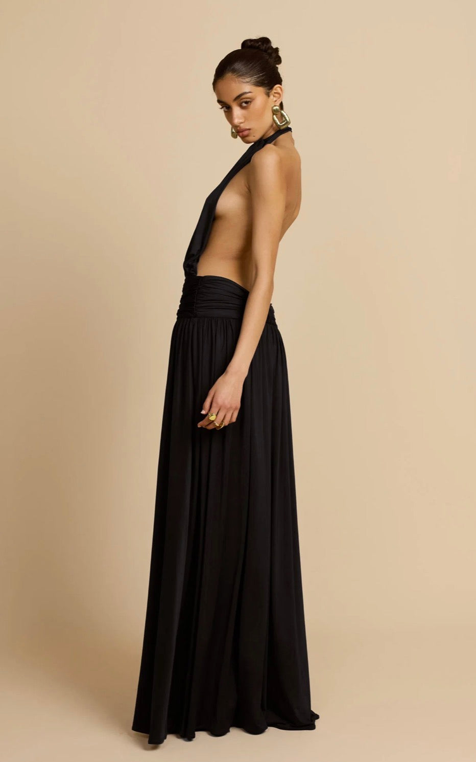A woman modeling a floor-length black halter gown with a side cutout and gathered waist, photographed against a plain warm beige studio background.