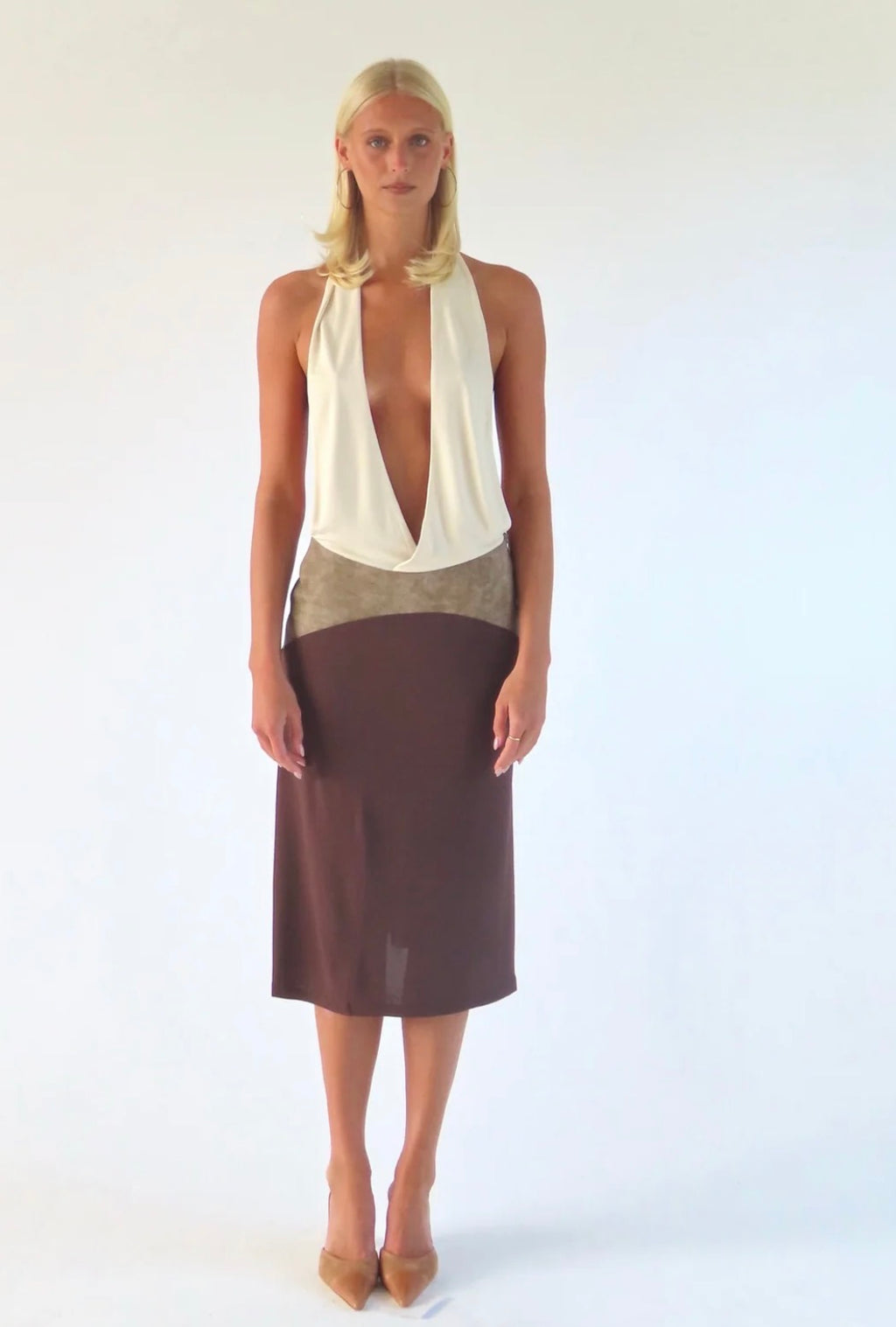 A blonde model stands against a white studio background wearing a color-block midi dress with a cream halter top, a taupe waistband, and a dark brown skirt, paired with tan heels.