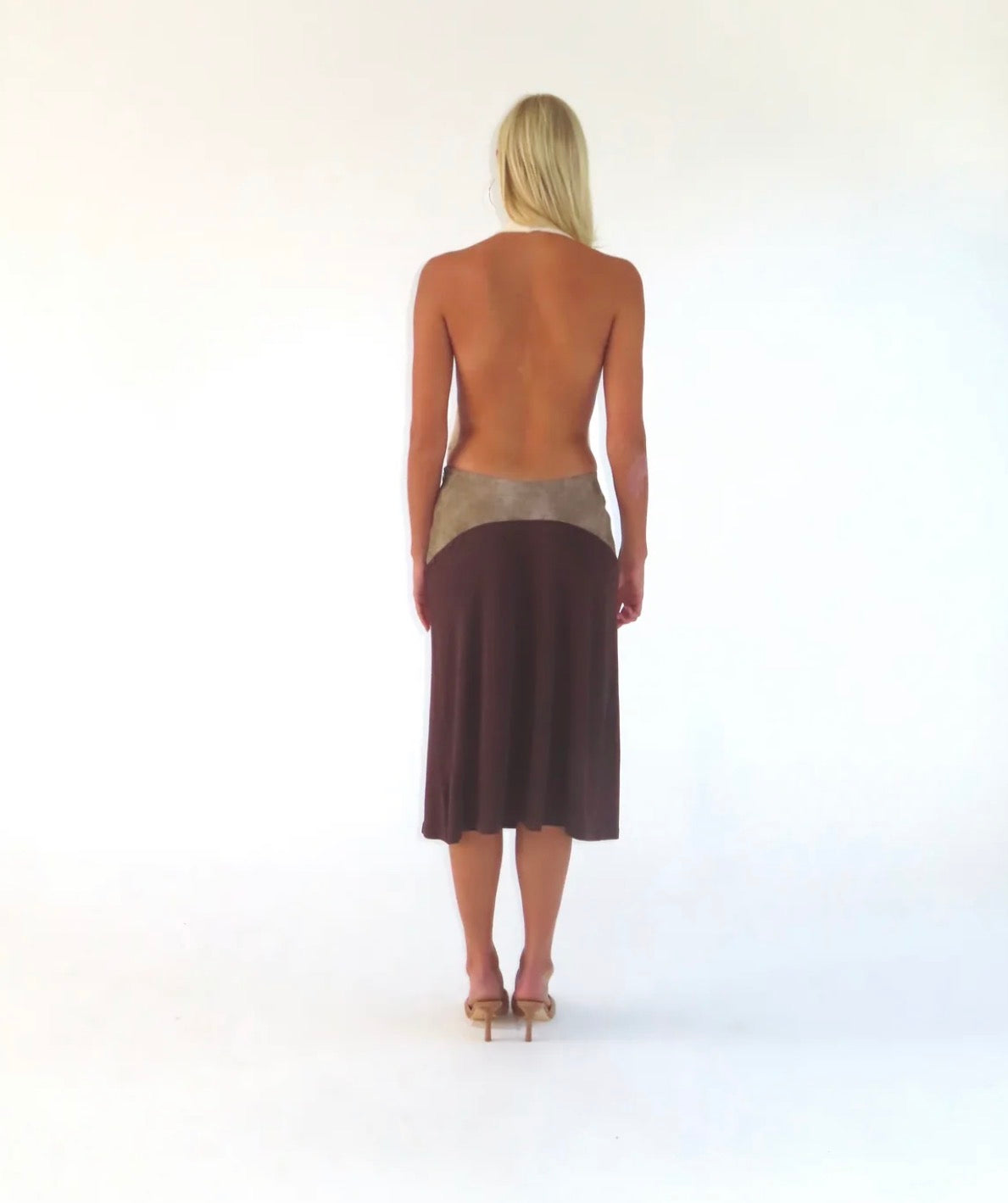 A woman stands with her back to the camera wearing a sleeveless, open-back midi dress with a metallic silver waistband and a dark brown skirt, set against a plain white studio background.