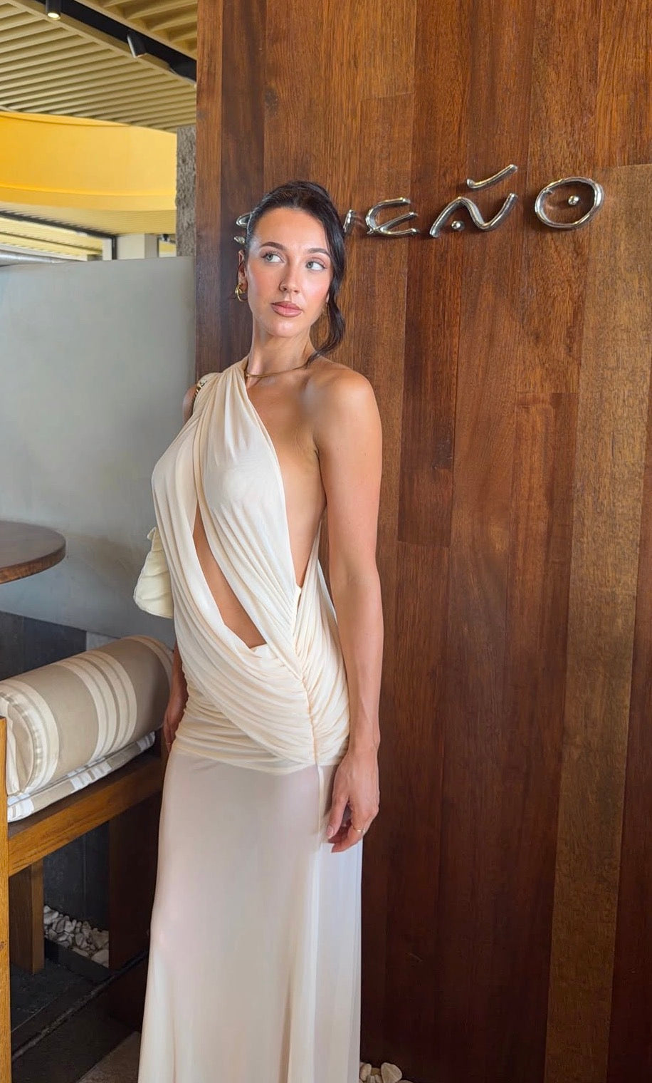 A woman wearing a cream-colored draped wrap dress with a cutout, standing in front of a wooden wall with a metal 'Teato' sign. - @alesha.beggs