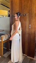 A woman wearing a cream-colored draped maxi dress with a deep back and long flowing fabric panel, standing in front of a dark wood-paneled wall with a 'SILVO' sign.
